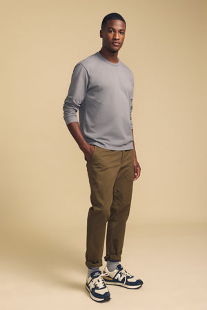 A man stands against a beige background with his hands in his pockets and a neutral expression, wearing the Velvet by Graham & Spencer ANTONIO TEE—a relaxed fit grey cotton jersey long sleeve—paired with khaki pants and black and white sneakers.