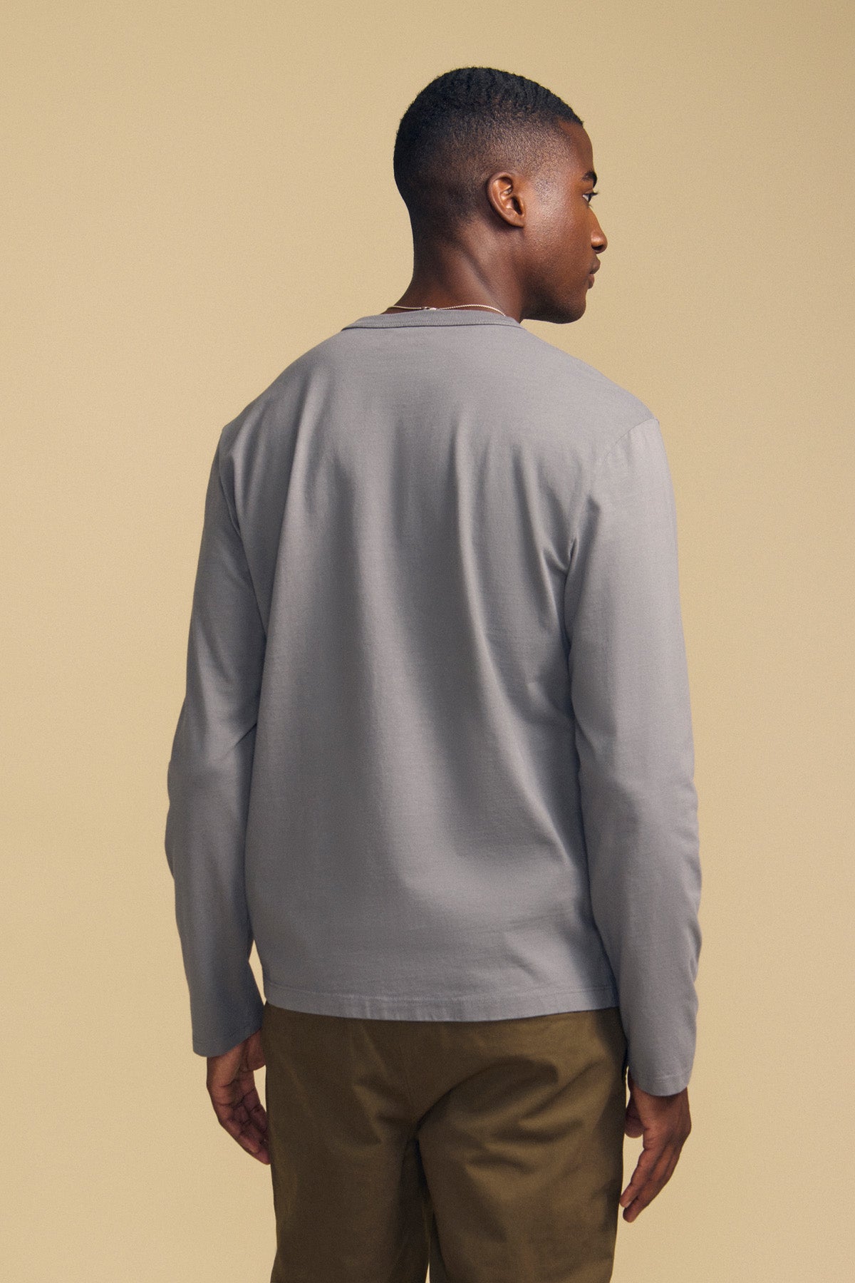 A person stands with their back to the camera, wearing the Velvet by Graham & Spencer ANTONIO TEE—a light gray cotton jersey long sleeve—paired with brown pants against a beige background.-42092437733569