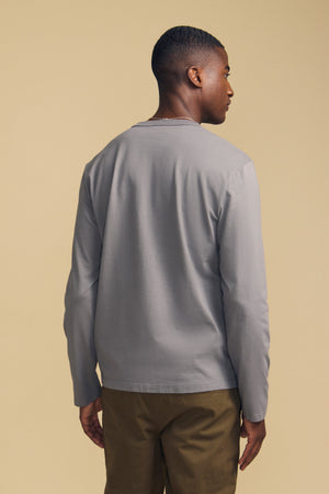 A person stands with their back to the camera, wearing the Velvet by Graham & Spencer ANTONIO TEE—a light gray cotton jersey long sleeve—paired with brown pants against a beige background.