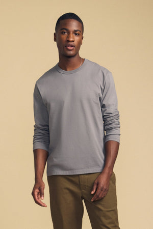 A person stands against a plain beige background, wearing the Velvet by Graham & Spencer ANTONIO TEE in gray cotton jersey with olive green pants, looking at the camera with a neutral expression.