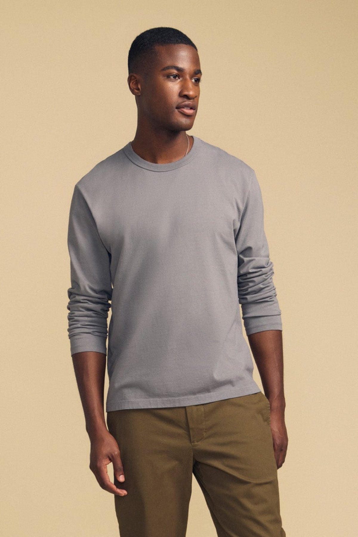 A man wearing the Velvet by Graham & Spencer ANTONIO TEE in light gray, styled in a relaxed fit with long sleeves and paired with olive pants, stands against a plain beige background, gazing slightly to the side.