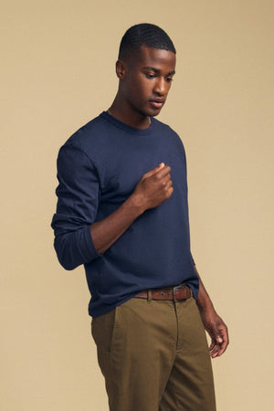 A person stands against a beige background, wearing the ANTONIO TEE from Velvet by Graham & Spencer—a navy cotton jersey long sleeve with a relaxed fit—paired with brown pants, a brown belt, and navy-and-white sneakers.