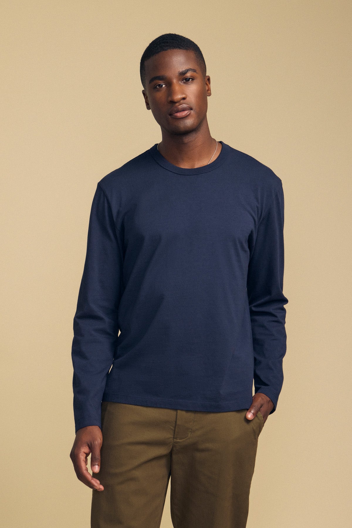 A person wearing the ANTONIO TEE by Velvet by Graham & Spencer—a navy cotton jersey long sleeve tee—paired with khaki pants stands against a plain beige background, looking at the camera with a neutral expression.