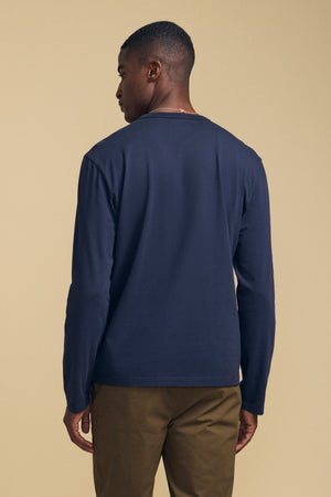 A person is standing with their back to the camera, wearing the Velvet by Graham & Spencer ANTONIO TEE in navy cotton jersey and olive green pants against a plain beige background.