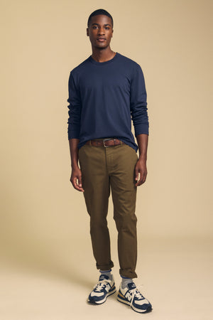 A man stands facing forward against a plain beige background, wearing the ANTONIO TEE by Velvet by Graham & Spencer—a relaxed fit navy cotton jersey long sleeve tee—paired with brown pants, a brown belt, and white and navy sneakers.