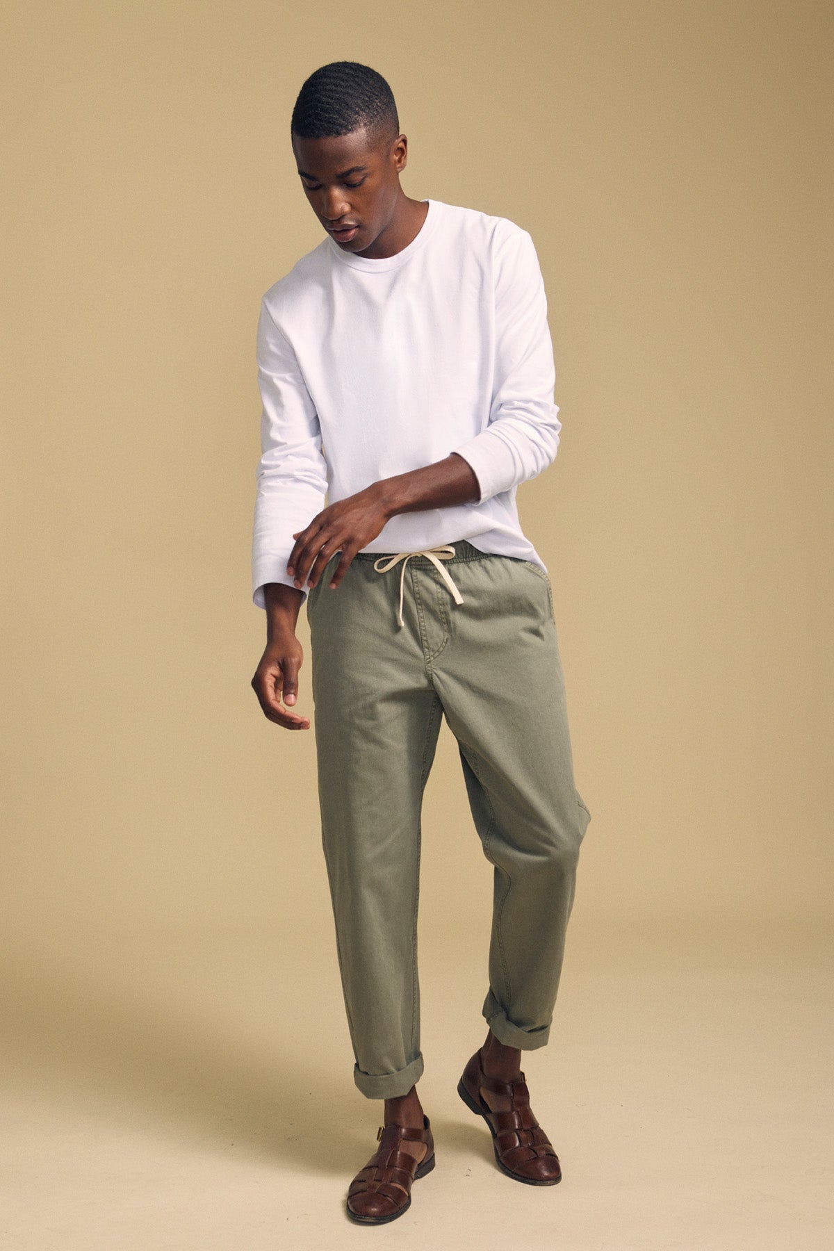 A person wearing the ANTONIO TEE by Velvet by Graham & Spencer, a white cotton jersey long sleeve top, pairs it with sage green relaxed fit drawstring pants and brown sandals, standing against a beige background.-41859586851009