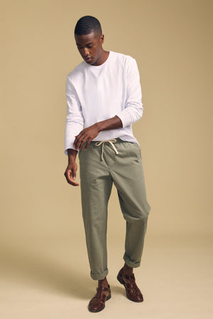 A person wearing the ANTONIO TEE by Velvet by Graham & Spencer, a white cotton jersey long sleeve top, pairs it with sage green relaxed fit drawstring pants and brown sandals, standing against a beige background.