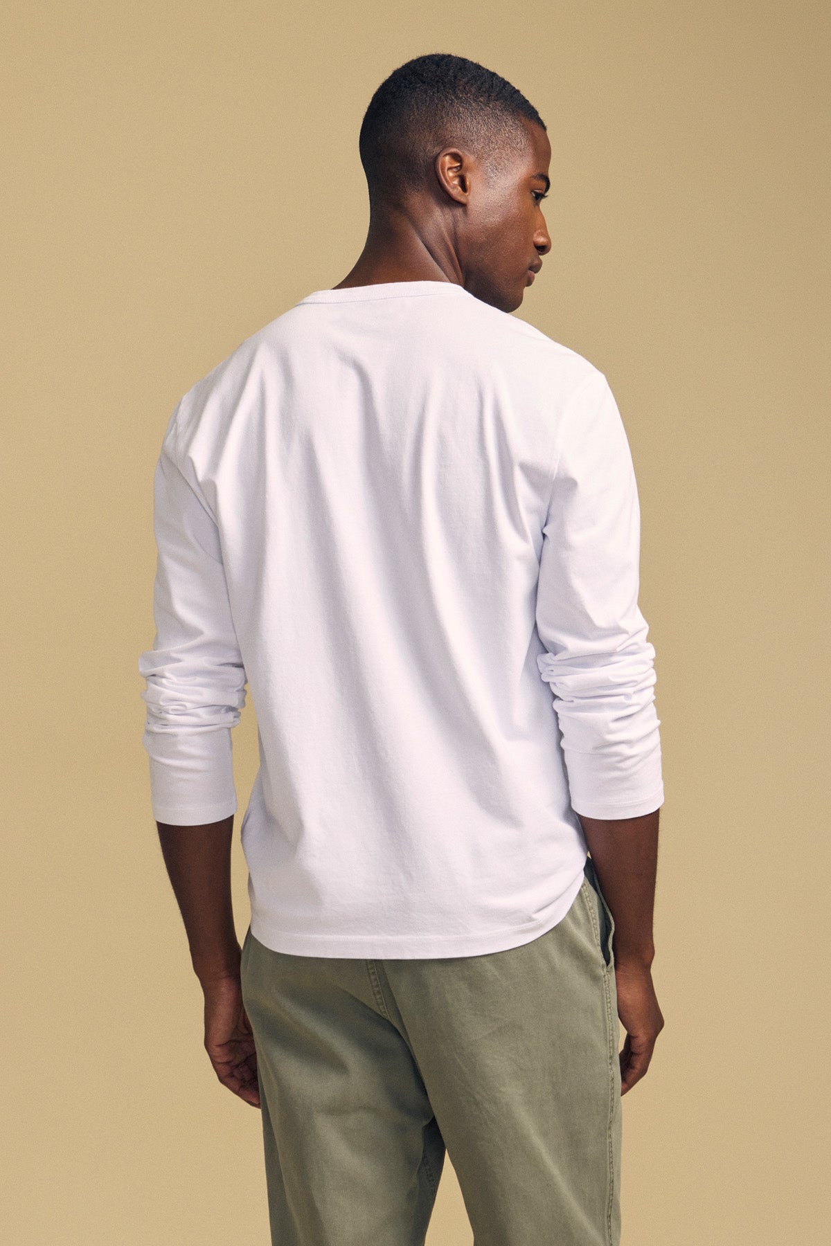 A man stands facing away against a plain beige background, wearing the Velvet by Graham & Spencer ANTONIO TEE in white, paired with light olive-green pants.