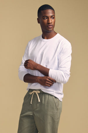 A man wearing the Velvet by Graham & Spencer ANTONIO TEE in white, styled with olive green drawstring pants, stands against a beige background and looks to the side.