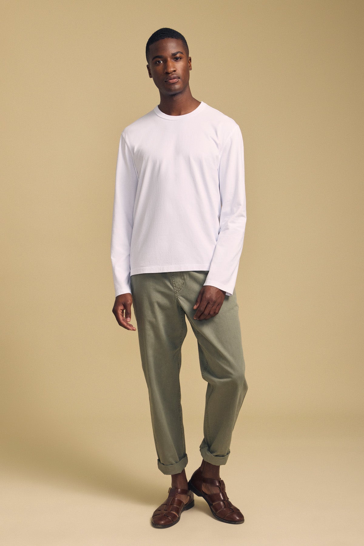 A man stands against a beige background wearing the ANTONIO TEE by Velvet by Graham & Spencer—a white long sleeve crafted from soft cotton jersey—paired with relaxed fit light olive green pants and brown shoes.