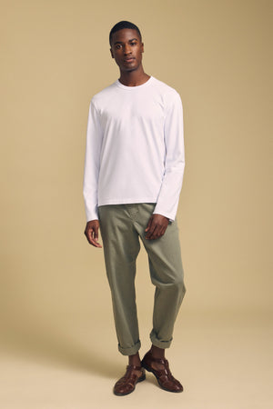 A man stands against a beige background wearing the ANTONIO TEE by Velvet by Graham & Spencer—a white long sleeve crafted from soft cotton jersey—paired with relaxed fit light olive green pants and brown shoes.