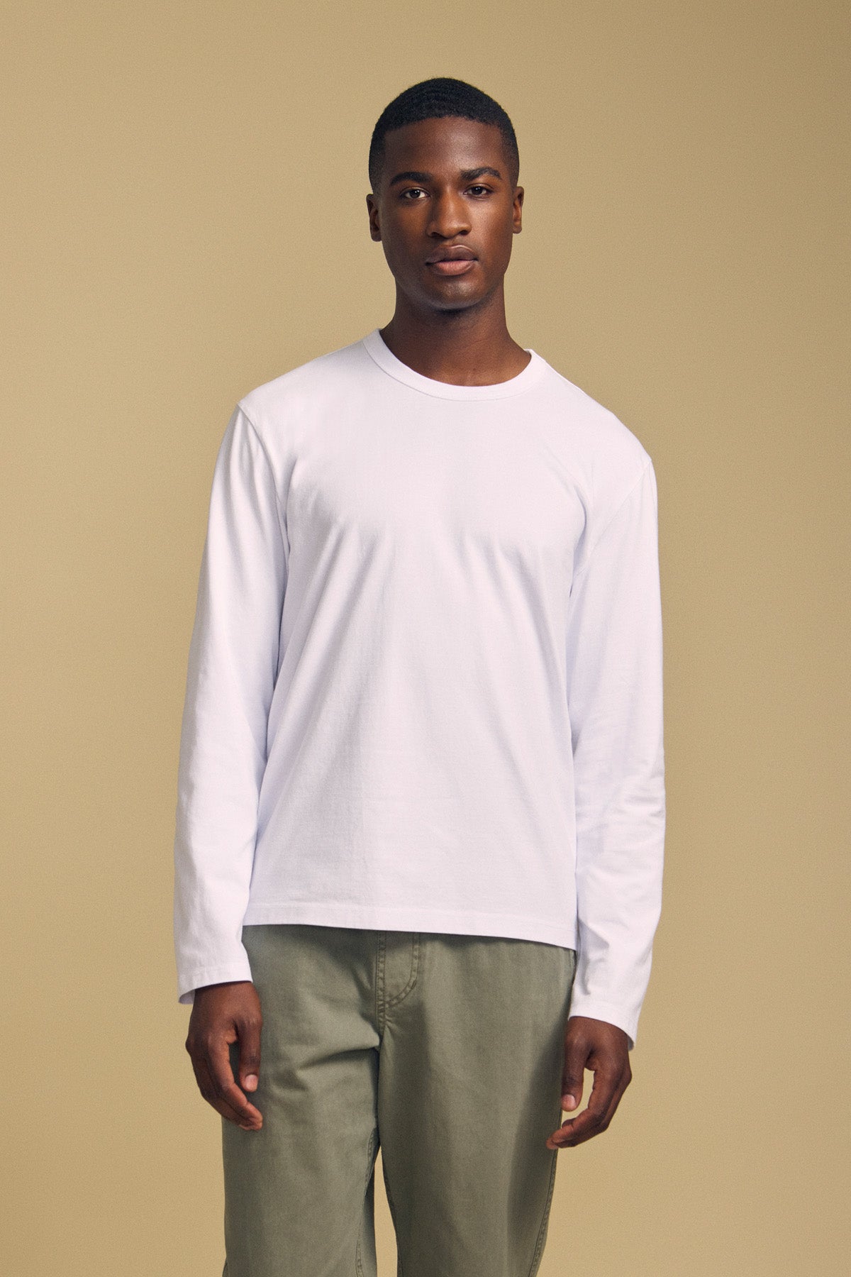 A man stands facing forward against a beige background, wearing the ANTONIO TEE by Velvet by Graham & Spencer—a relaxed fit, long sleeve shirt crafted from soft cotton jersey—paired with olive green pants.