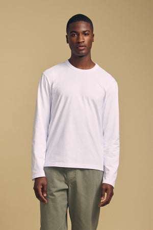 A man stands facing forward against a beige background, wearing the ANTONIO TEE by Velvet by Graham & Spencer—a relaxed fit, long sleeve shirt crafted from soft cotton jersey—paired with olive green pants.