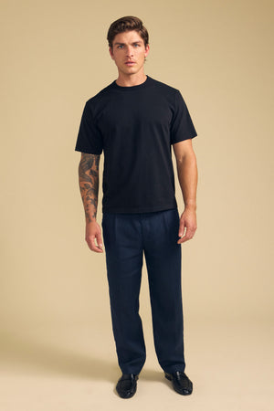 A man faces forward against a beige background, wearing the WELLS TEE by Velvet by Graham & Spencer in black Pima cotton jersey, dark blue pants, and black shoes. He shows a tattoo sleeve on his right arm. Ideal for all-day comfort.
