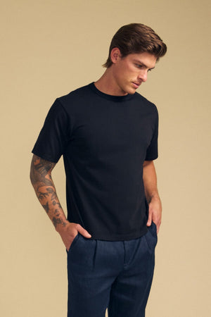 A person with tattoos on their left arm wears the Velvet by Graham & Spencer WELLS TEE, a relaxed fit plain black Pima cotton jersey, paired with dark pants and hands in pockets against a beige background—ideal for all-day wear.