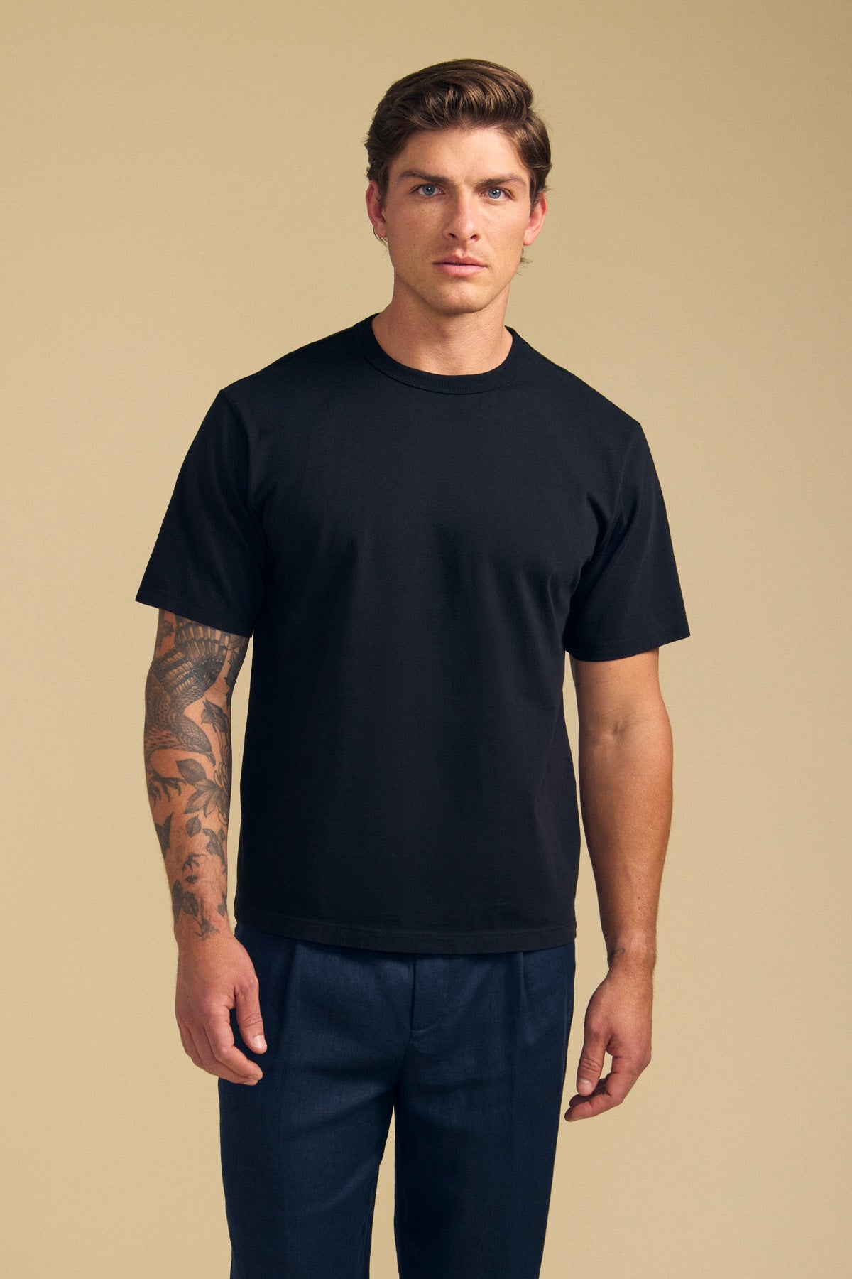 A man with short light brown hair and a tattoo on his left arm wears the Velvet by Graham & Spencer WELLS TEE in relaxed fit Pima cotton jersey and dark pants, standing against a beige background—ideal for all-day wear.