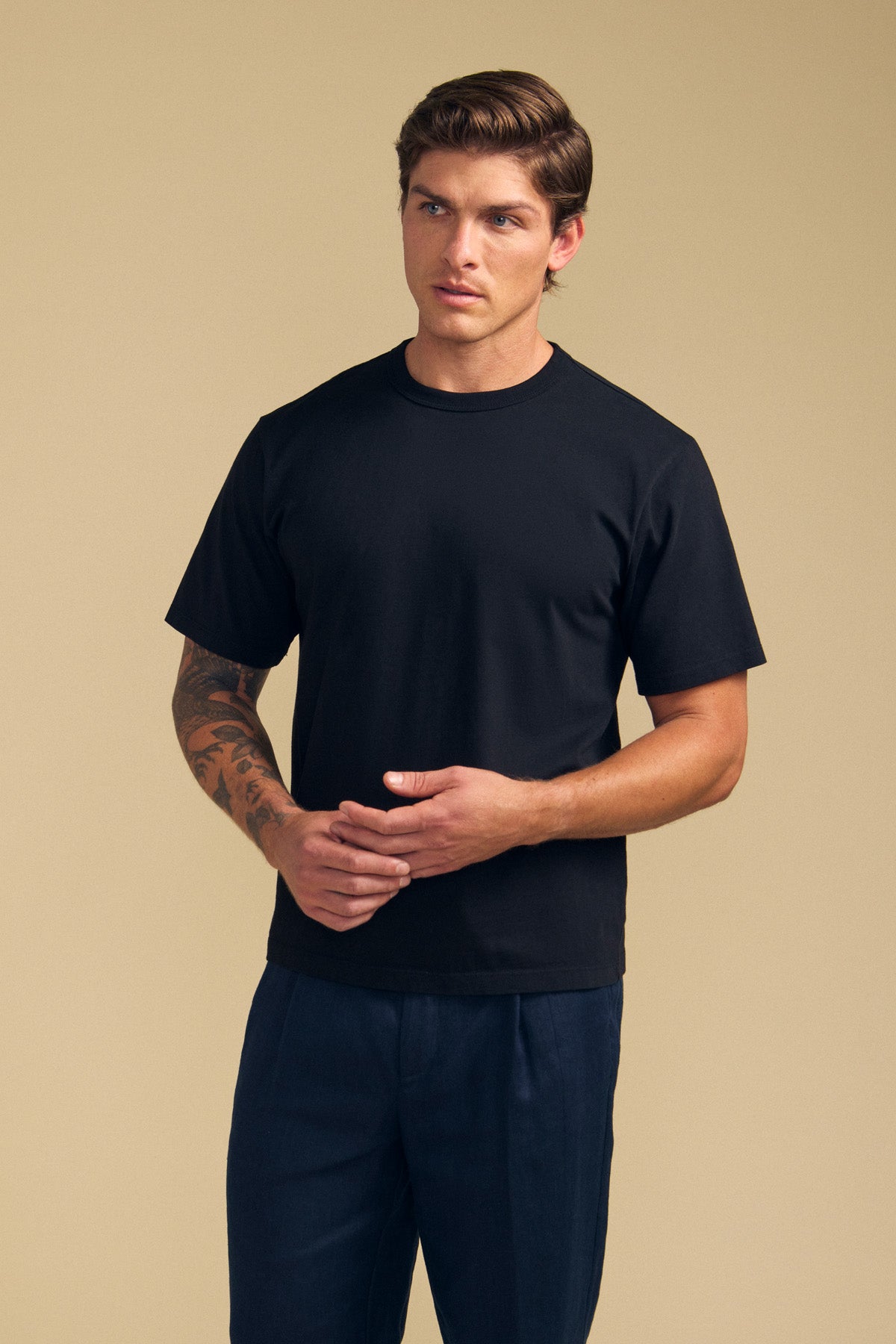 A man with short brown hair, wearing the WELLS TEE by Velvet by Graham & Spencer and dark pants, stands against a beige background with his hands clasped.