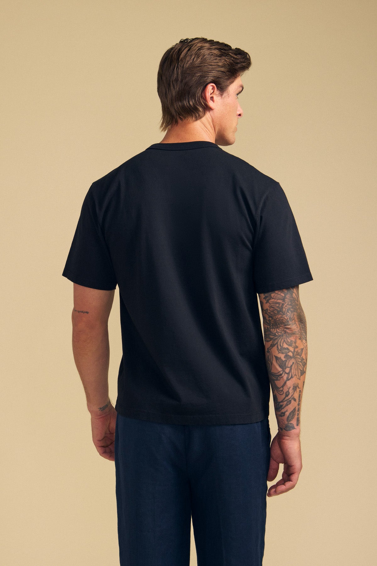 A man stands facing away against a beige background, wearing the Velvet by Graham & Spencer WELLS TEE in black Pima cotton jersey, paired with dark pants and showing tattoos on his right arm.