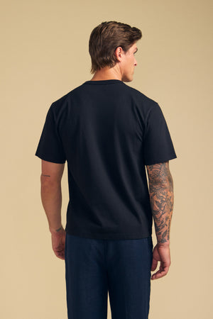 A man stands facing away against a beige background, wearing the Velvet by Graham & Spencer WELLS TEE in black Pima cotton jersey, paired with dark pants and showing tattoos on his right arm.