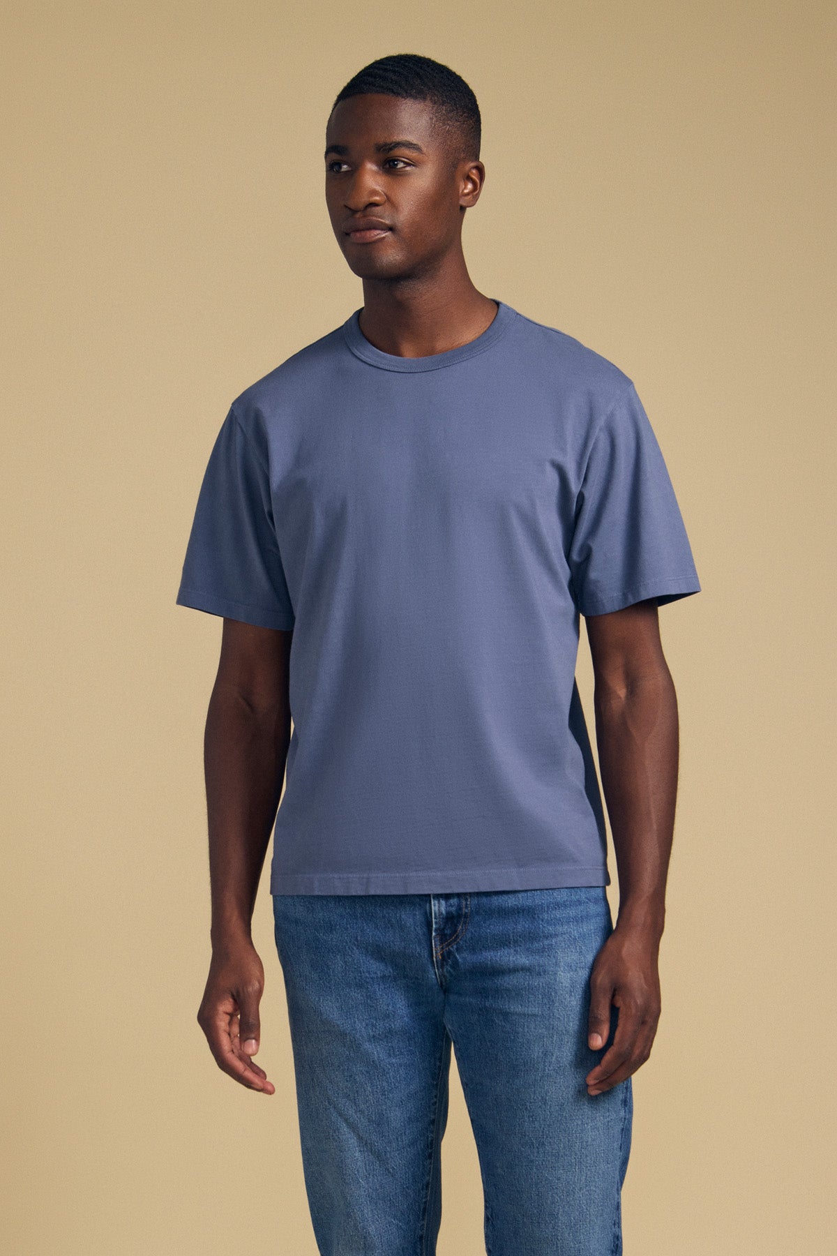 A man stands against a plain beige background, wearing the WELLS TEE by Velvet by Graham & Spencer—a relaxed fit blue t-shirt crafted from soft Pima cotton jersey—paired with blue jeans. He gazes slightly to the side with a neutral expression, while the crew neckline enhances his classic and understated style.