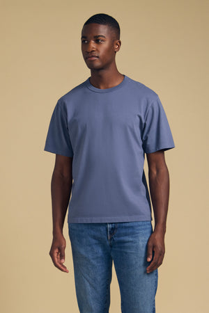 A man stands against a plain beige background, wearing the WELLS TEE by Velvet by Graham & Spencer—a relaxed fit blue t-shirt crafted from soft Pima cotton jersey—paired with blue jeans. He gazes slightly to the side with a neutral expression, while the crew neckline enhances his classic and understated style.