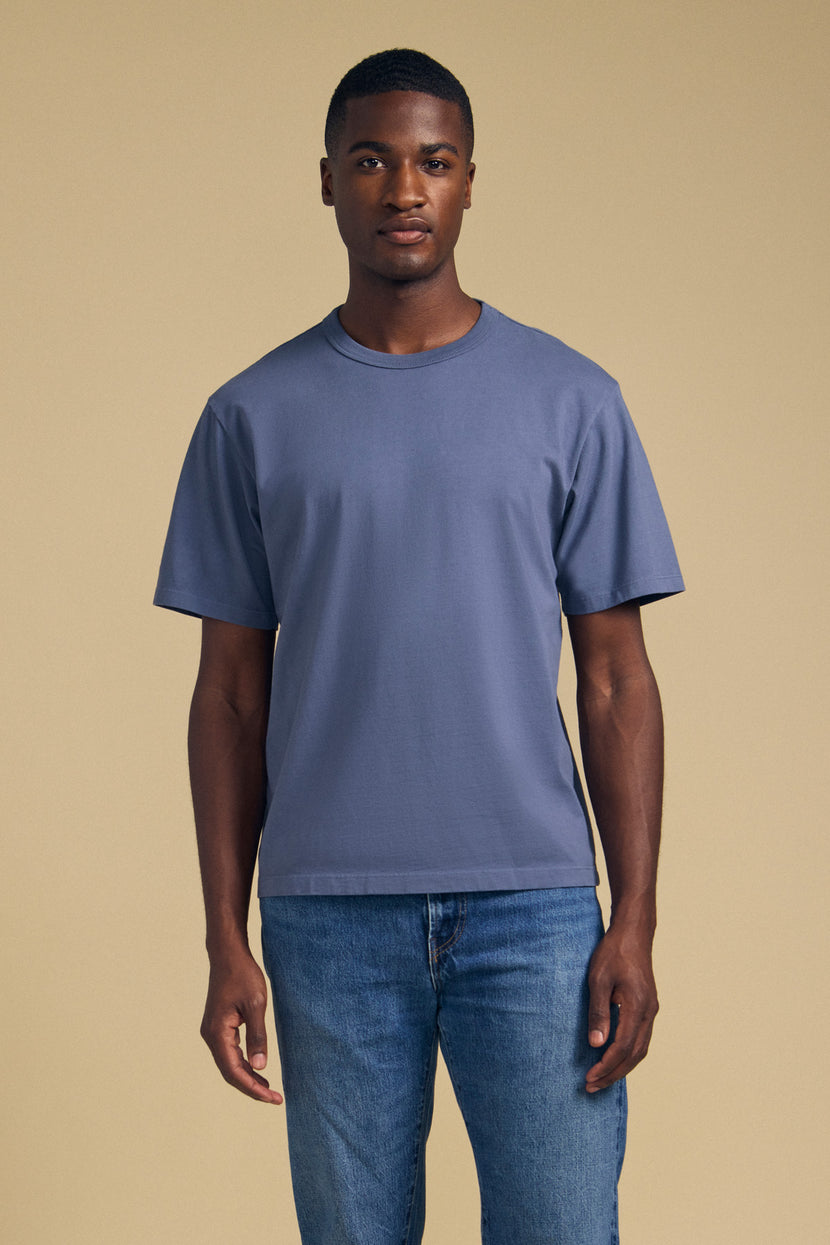 A man stands facing forward wearing the Velvet by Graham & Spencer WELLS TEE—a blue short-sleeve t-shirt featuring a classic crew neckline and relaxed fit—paired with blue jeans against a plain beige background.