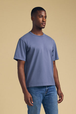 A person stands against a beige background, wearing the Velvet by Graham & Spencer WELLS TEE—a faded blue short-sleeve t-shirt made from soft Pima cotton jersey—paired with blue jeans, facing slightly to the right. The shirt’s relaxed fit offers comfortable all-day wear.