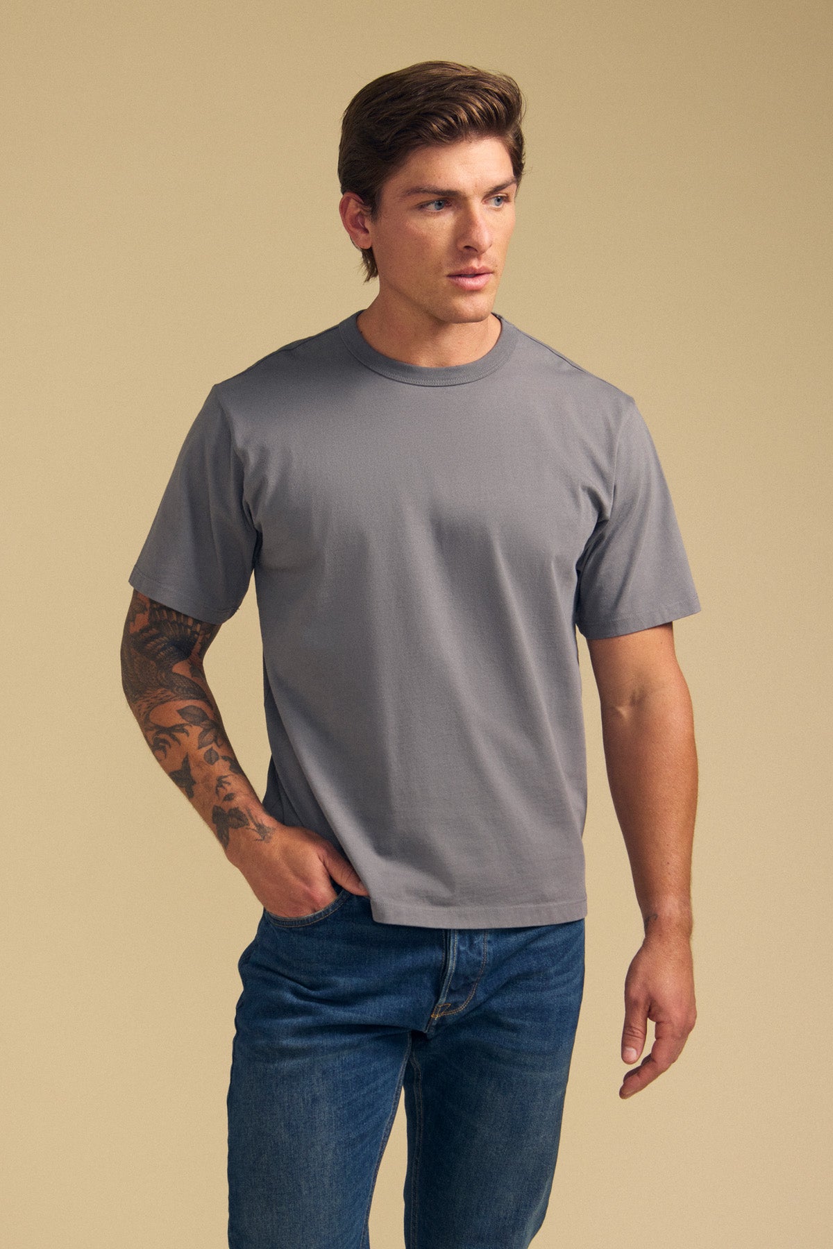 A man with a tattooed left arm wears the Velvet by Graham & Spencer WELLS TEE, a relaxed fit plain gray Pima cotton jersey, paired with blue jeans, standing against a beige background.