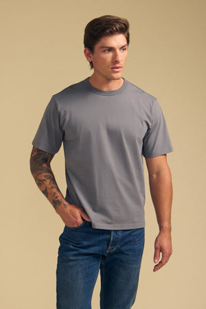 A man with a tattooed left arm wears the Velvet by Graham & Spencer WELLS TEE, a relaxed fit plain gray Pima cotton jersey, paired with blue jeans, standing against a beige background.