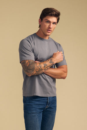 A man with brown hair and a tattooed right arm wears the Velvet by Graham & Spencer WELLS TEE in gray Pima cotton jersey and blue jeans, posing against a beige background—ideal for all-day comfort.