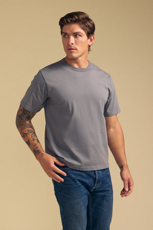 A man with short brown hair and tattoos on his left arm wears the Velvet by Graham & Spencer WELLS TEE in plain gray Pima cotton jersey and blue jeans, standing against a beige background.