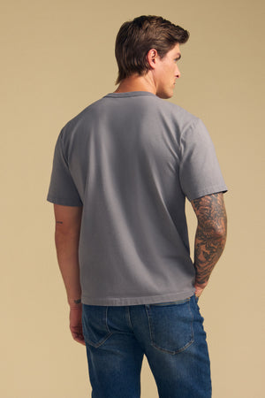 A man with short brown hair, wearing the WELLS TEE by Velvet by Graham & Spencer in gray Pima cotton jersey and blue jeans, stands with his back to the camera against a plain beige background.