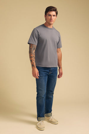 A man stands against a beige background wearing the WELLS TEE in gray Pima cotton jersey by Velvet by Graham & Spencer, paired with blue jeans and light-colored sneakers.