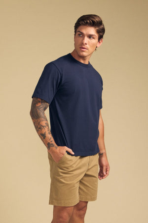 A man with a tattooed arm wears the Velvet by Graham & Spencer WELLS TEE, a relaxed fit navy Pima cotton jersey t-shirt, paired with tan shorts. He stands against a beige background with one hand in his pocket.