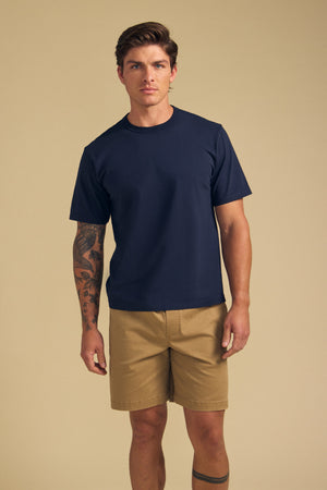 A man with a sleeve tattoo stands against a beige background wearing the WELLS TEE by Velvet by Graham & Spencer—a navy blue Pima cotton jersey crewneck t-shirt—paired with khaki shorts.