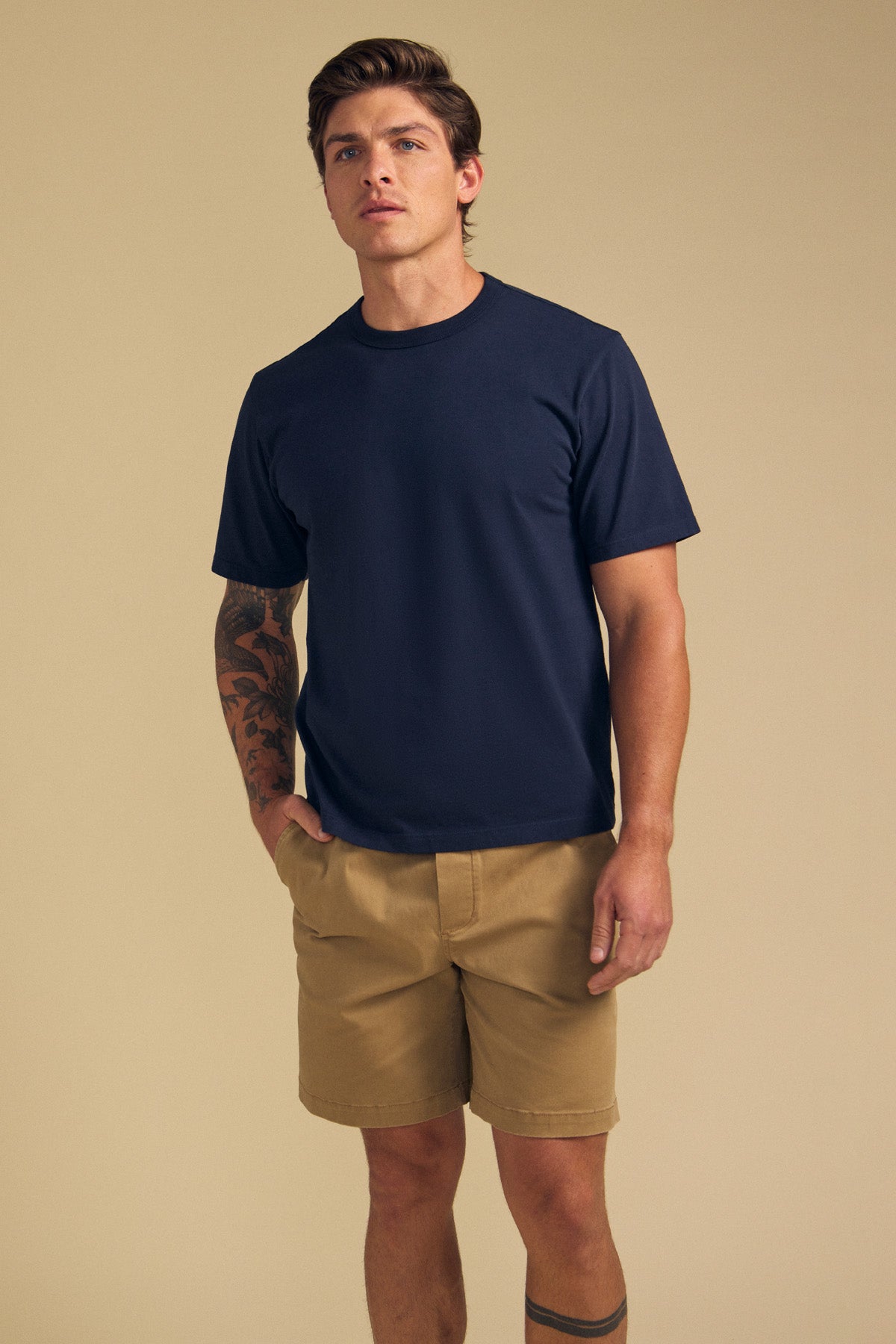 A man poses against a beige background in the Velvet by Graham & Spencer WELLS TEE, a navy blue Pima cotton jersey crewneck, paired with khaki shorts. Tattoos on his left forearm and right leg add to his relaxed style.