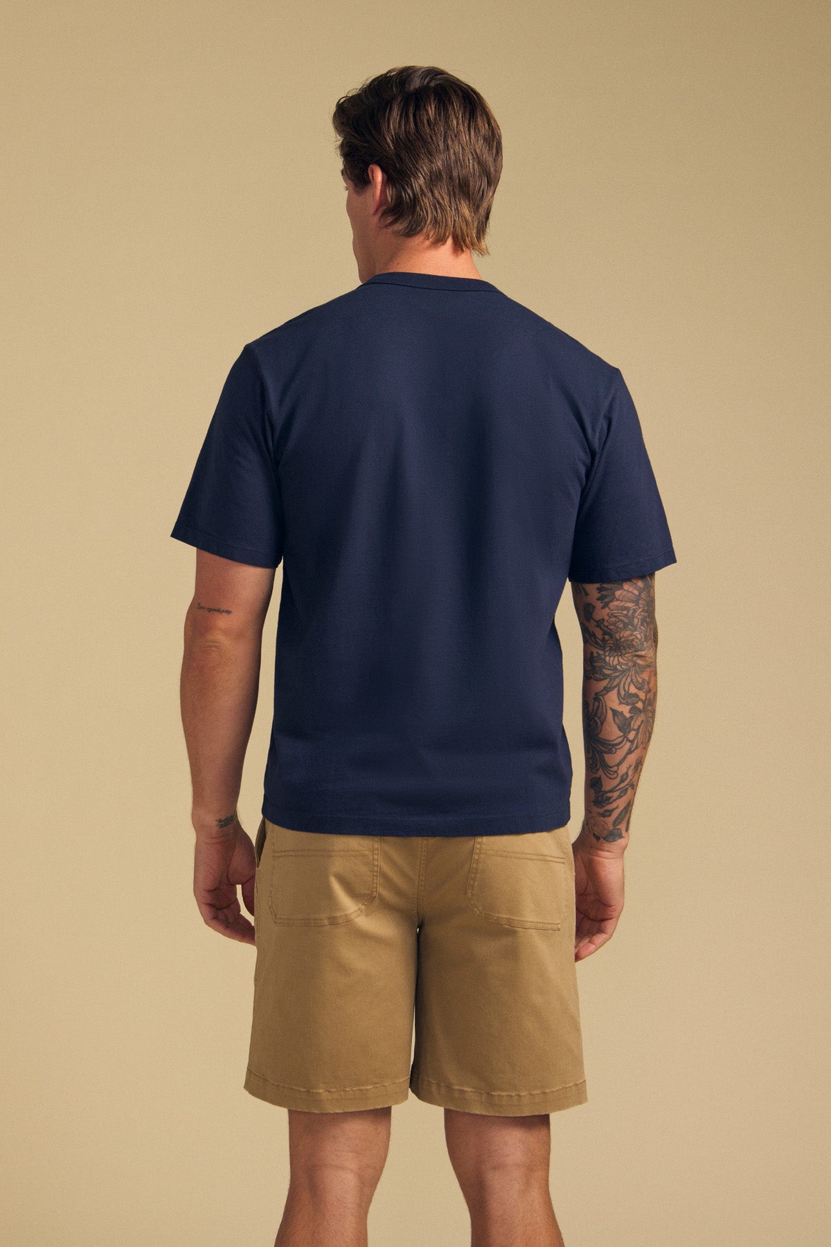 A man faces away, wearing the WELLS TEE by Velvet by Graham & Spencer—navy blue Pima cotton jersey with a relaxed fit—and khaki shorts. A tattoo sleeve is visible on his right arm. The background is plain beige.