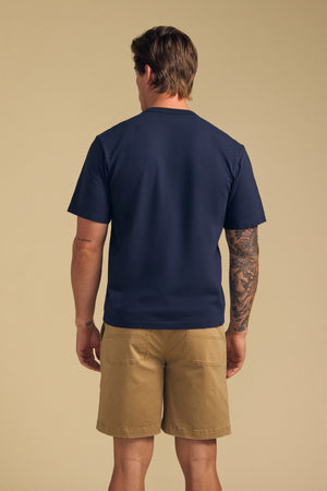 A man faces away, wearing the WELLS TEE by Velvet by Graham & Spencer—navy blue Pima cotton jersey with a relaxed fit—and khaki shorts. A tattoo sleeve is visible on his right arm. The background is plain beige.