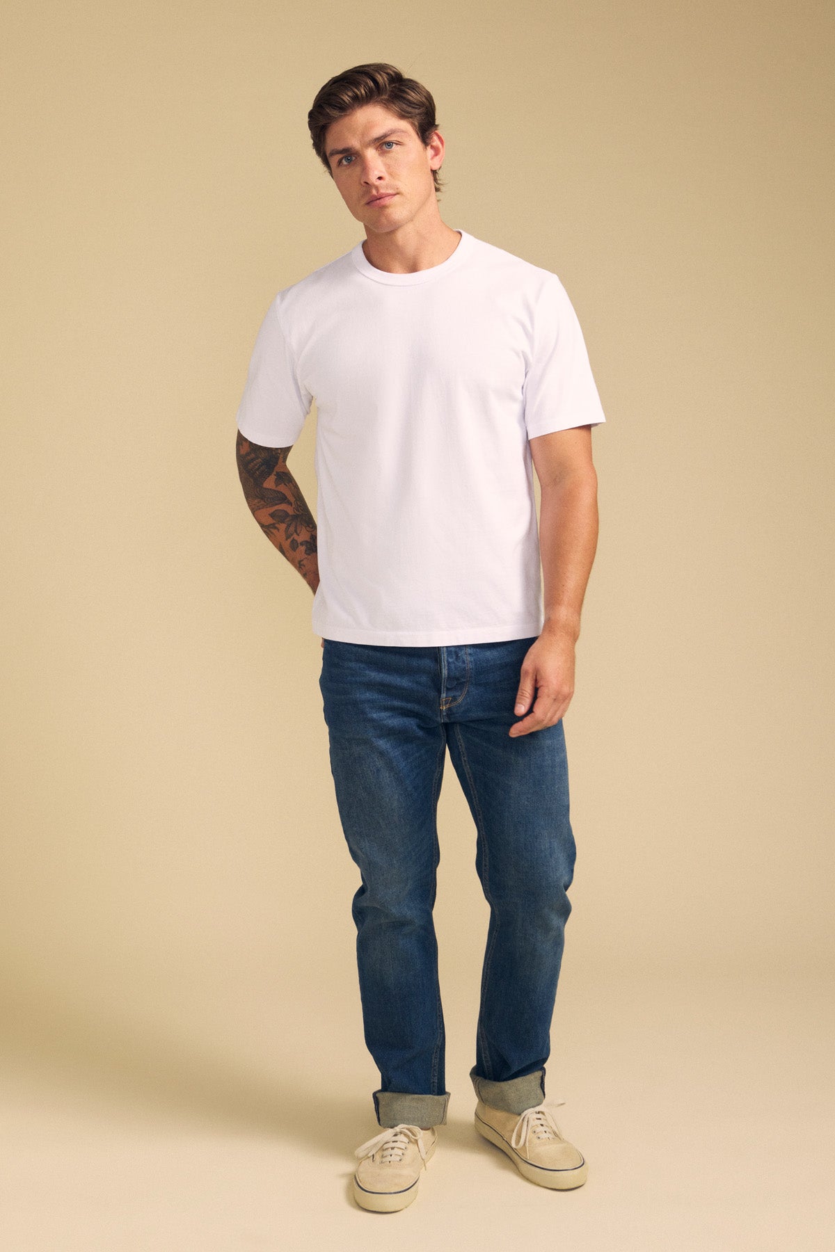 A man stands against a beige background, wearing the WELLS TEE by Velvet by Graham & Spencer—a white Pima cotton tee in a relaxed fit—paired with blue jeans and beige shoes. A tattoo is visible on one arm.-41774610776257