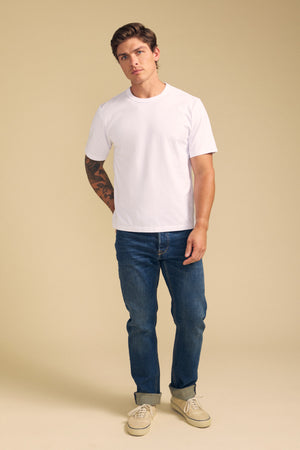 A man stands against a beige background, wearing the WELLS TEE by Velvet by Graham & Spencer—a white Pima cotton tee in a relaxed fit—paired with blue jeans and beige shoes. A tattoo is visible on one arm.