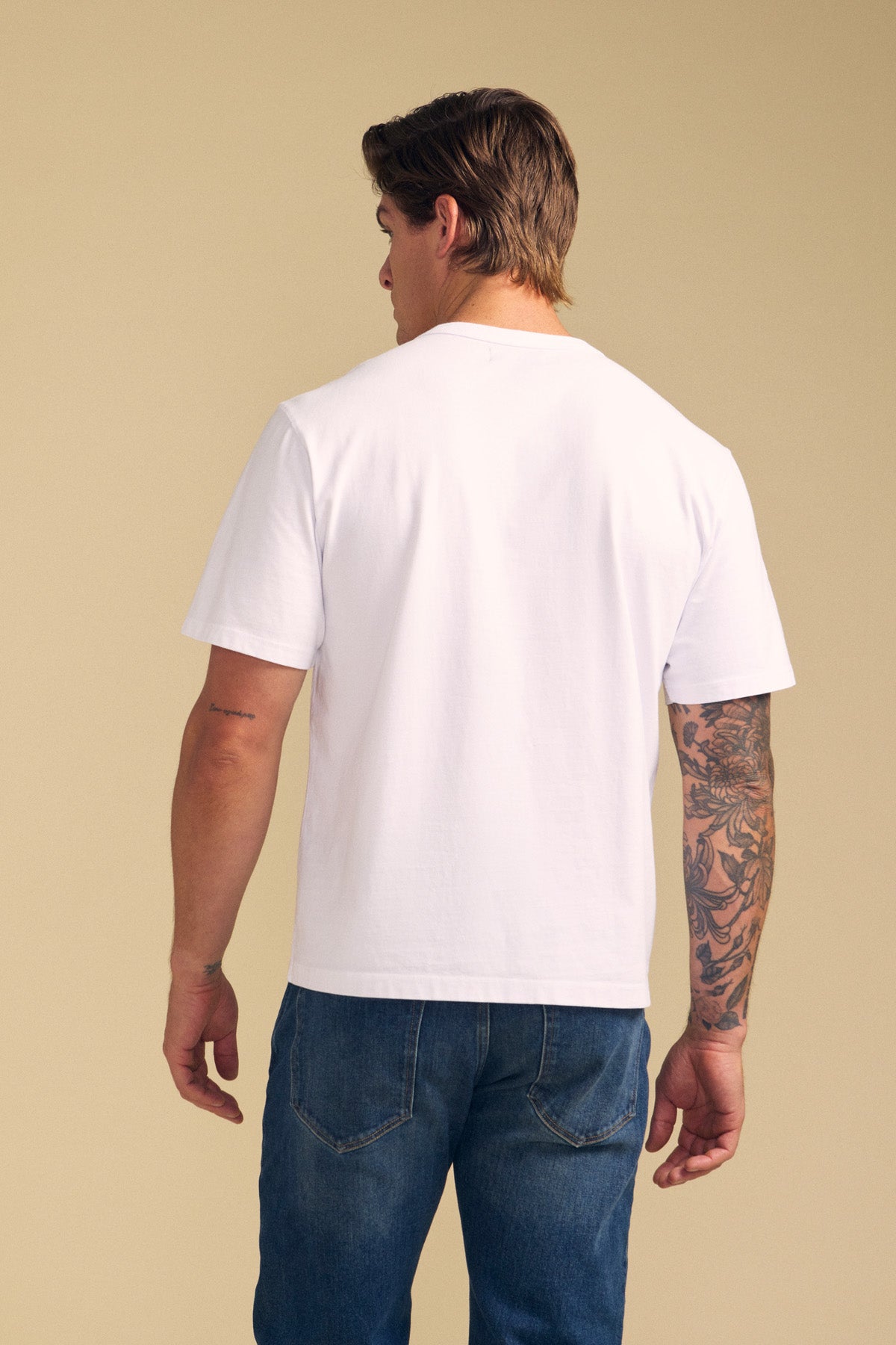 A person stands facing away, wearing the WELLS TEE by Velvet by Graham & Spencer—a relaxed fit plain white Pima cotton tee—paired with blue jeans. One arm is tattooed. The background is a neutral beige color.