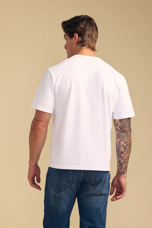 A person stands facing away, wearing the WELLS TEE by Velvet by Graham & Spencer—a relaxed fit plain white Pima cotton tee—paired with blue jeans. One arm is tattooed. The background is a neutral beige color.
