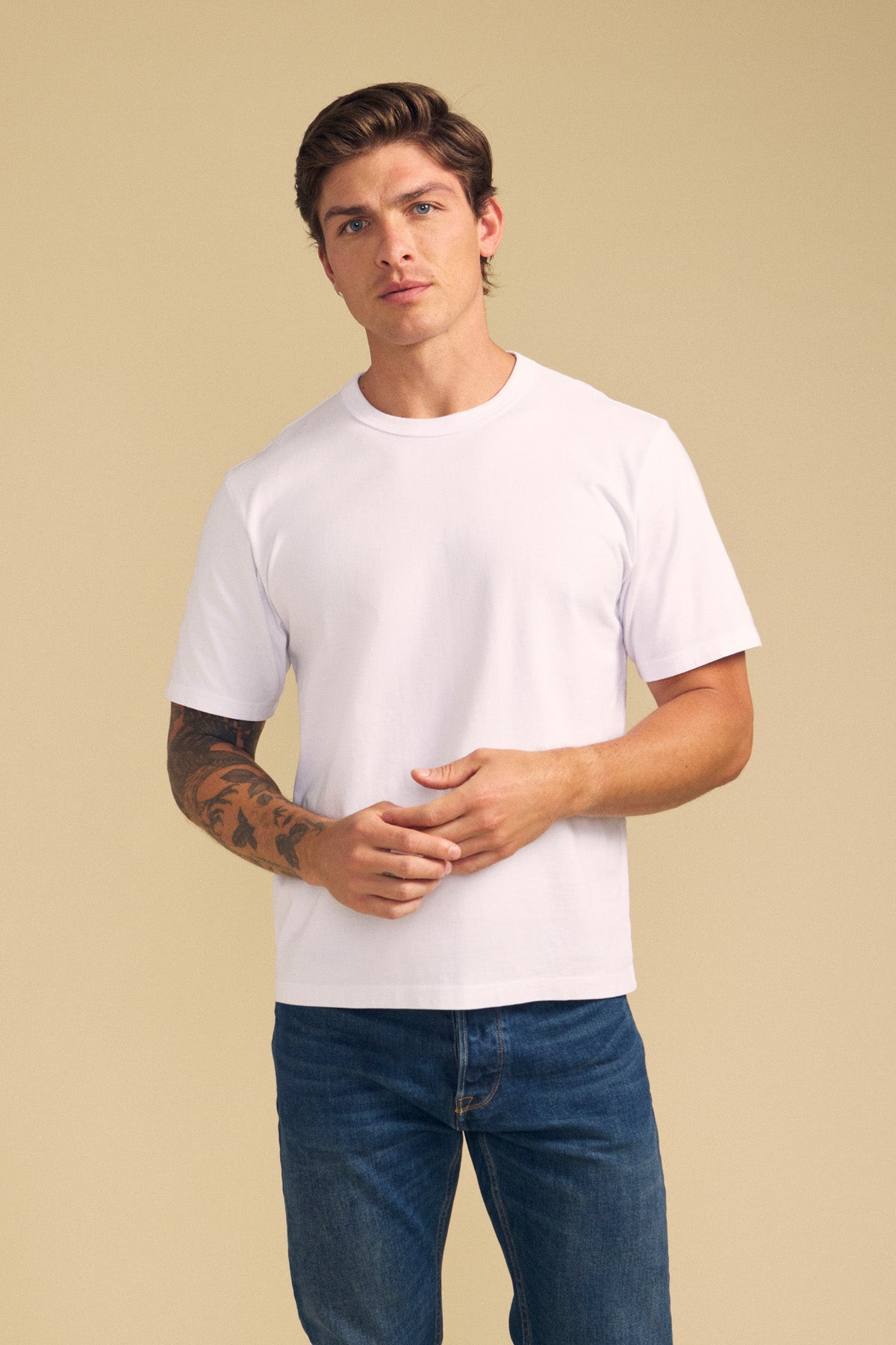 A man with short brown hair and a tattoo on his left arm wears the WELLS TEE by Velvet by Graham & Spencer—a plain white t-shirt with a classic crew neckline—paired with blue jeans, standing against a beige background.-41774610710721