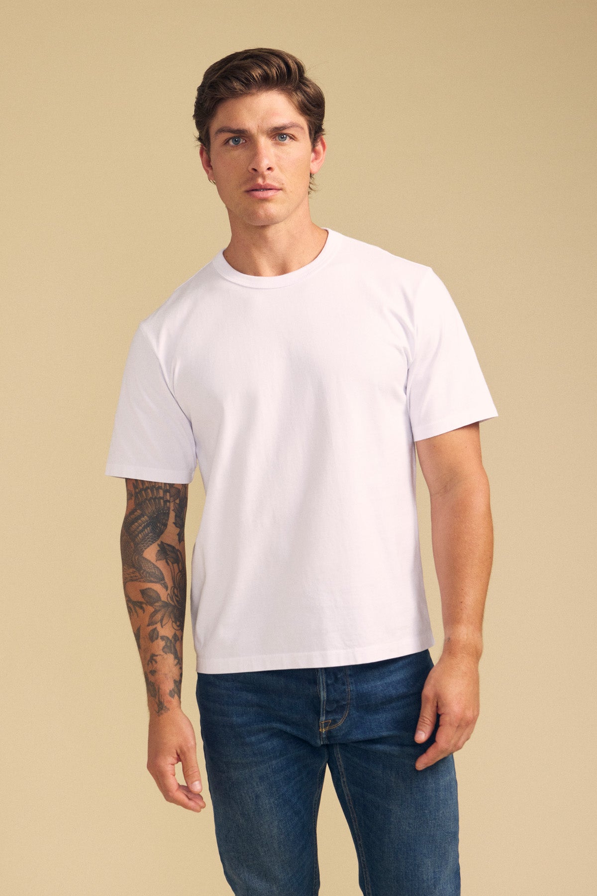 A man with short brown hair and a tattooed left arm wears the WELLS TEE—a relaxed fit, plain white Pima cotton t-shirt by Velvet by Graham & Spencer—paired with blue jeans, standing against a beige background.