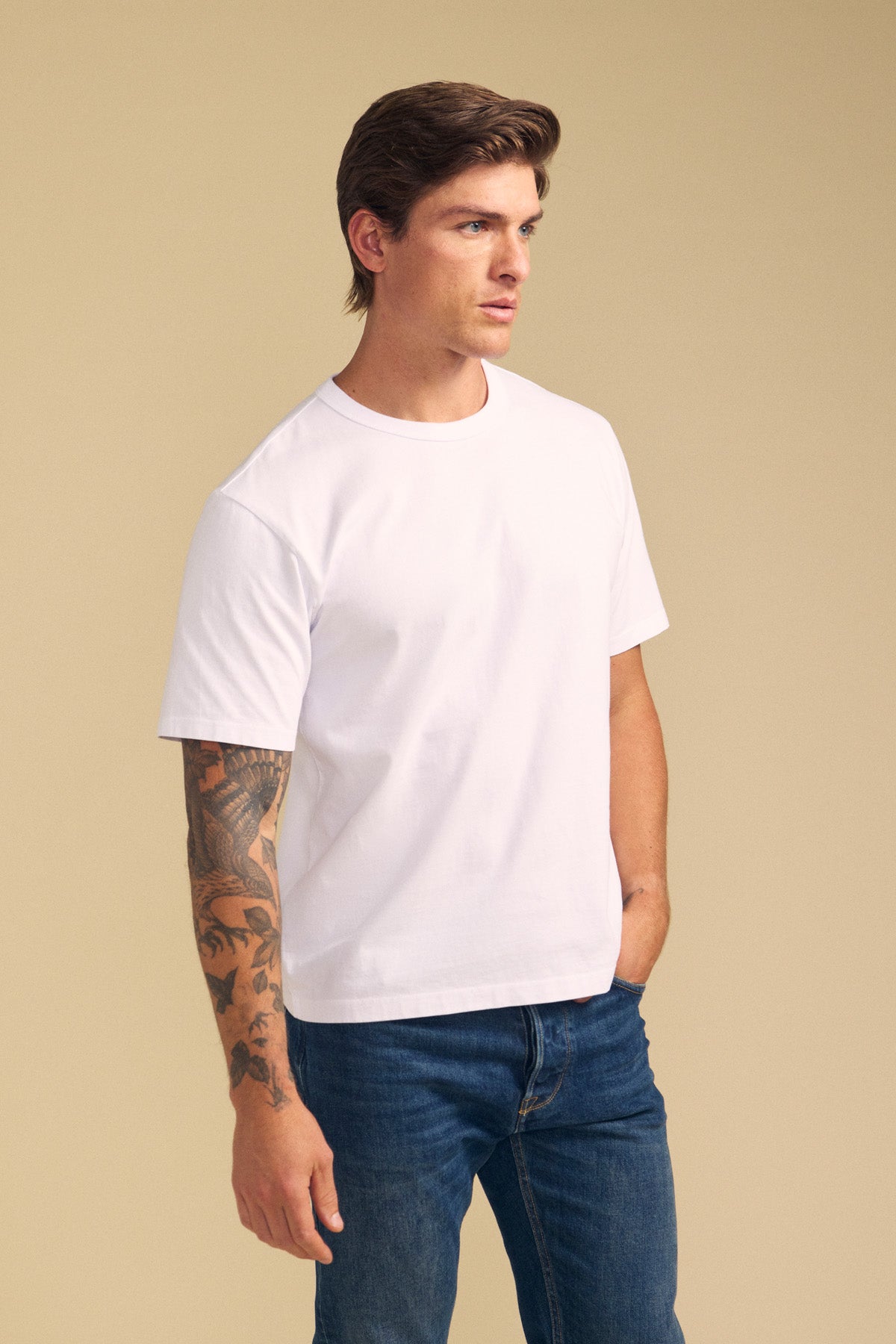 A man with brown hair and a tattooed arm wears the relaxed fit WELLS TEE in plain white Pima cotton by Velvet by Graham & Spencer, paired with blue jeans, standing against a beige background.