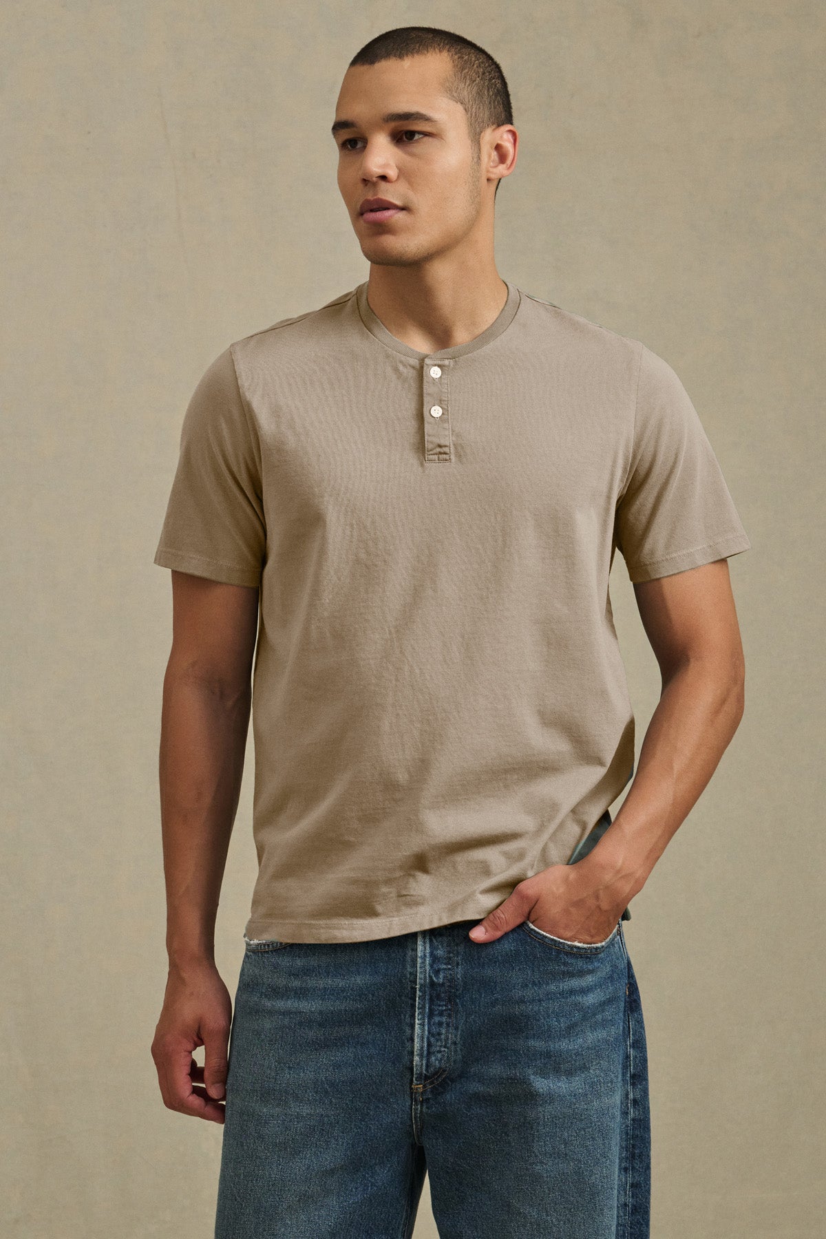 A man stands against a neutral background with one hand in his pocket, wearing the FELIX HENLEY by Velvet by Graham & Spencer in beige Pima cotton and blue jeans.