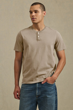 A man stands against a neutral background with one hand in his pocket, wearing the FELIX HENLEY by Velvet by Graham & Spencer in beige Pima cotton and blue jeans.