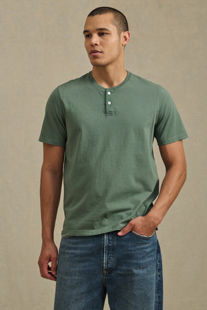 A man wearing the Velvet by Graham & Spencer FELIX HENLEY in green and blue jeans stands against a plain beige background.