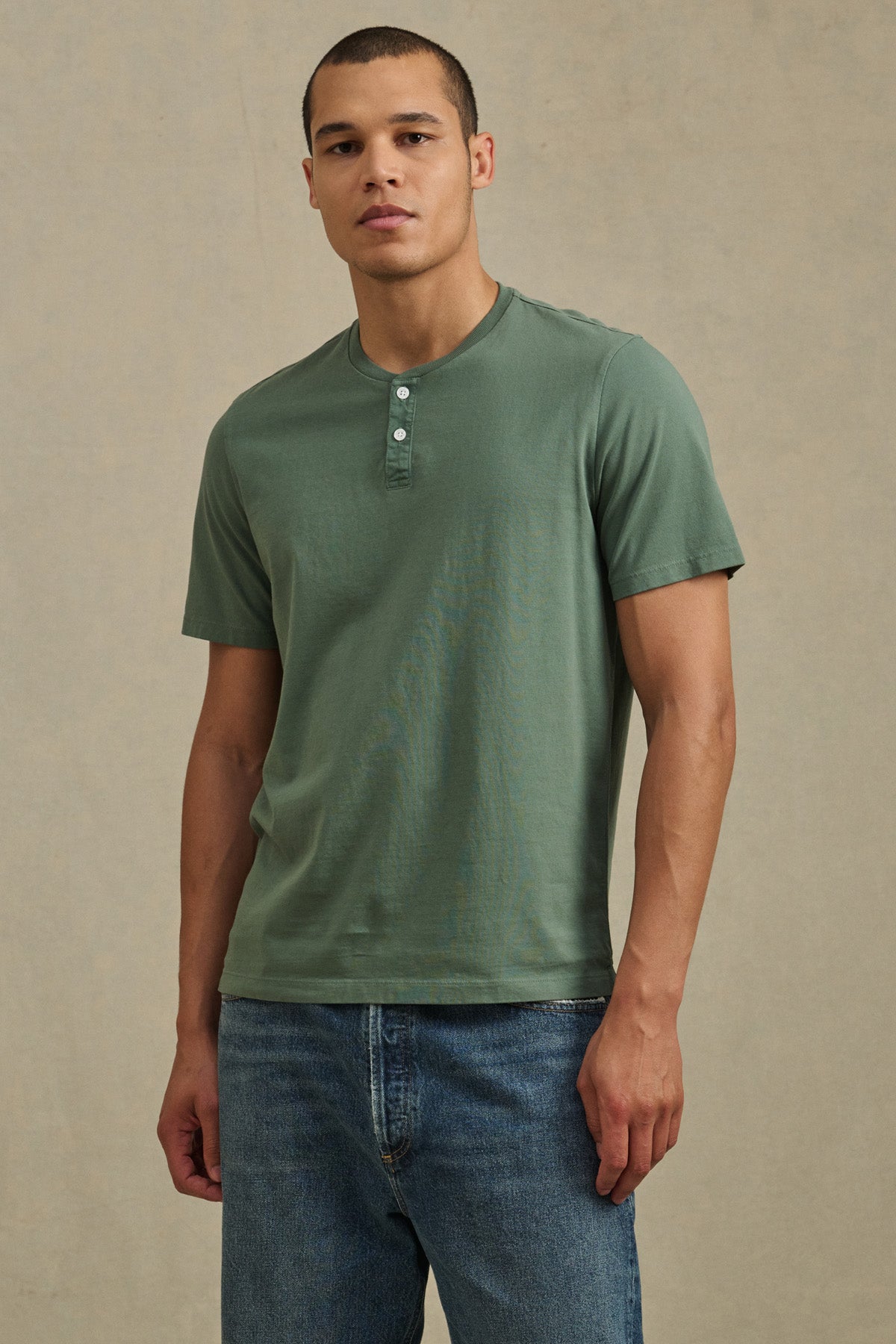 A man stands against a neutral background wearing the FELIX HENLEY by Velvet by Graham & Spencer in green, paired with blue jeans.