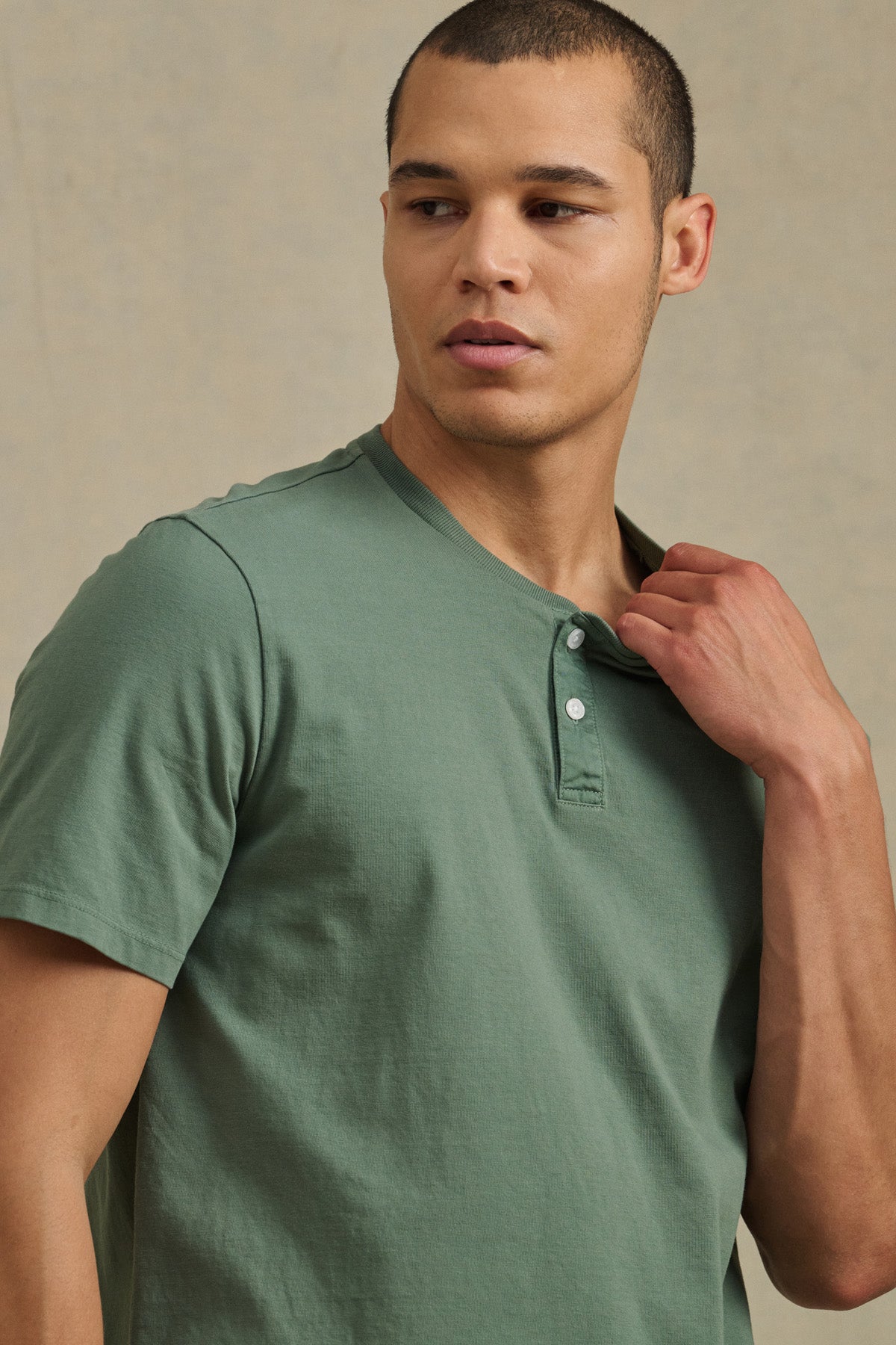 A man wearing the FELIX HENLEY by Velvet by Graham & Spencer—a short sleeve green Pima cotton henley—looks to the side, holding the collar with one hand against a neutral background.-41479917142209